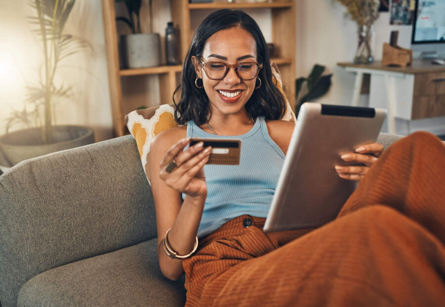 A young woman laying down on the couch smiles while holding her credit card and using her tablet.