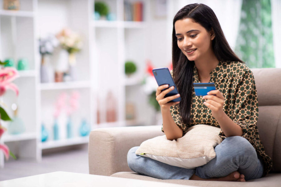 A young women smiles as she is sitting on the couch while holding her phone and credit card.