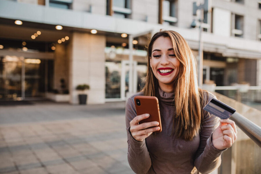 A young woman smiles as she holds her phone while holding her credit card in her hand.