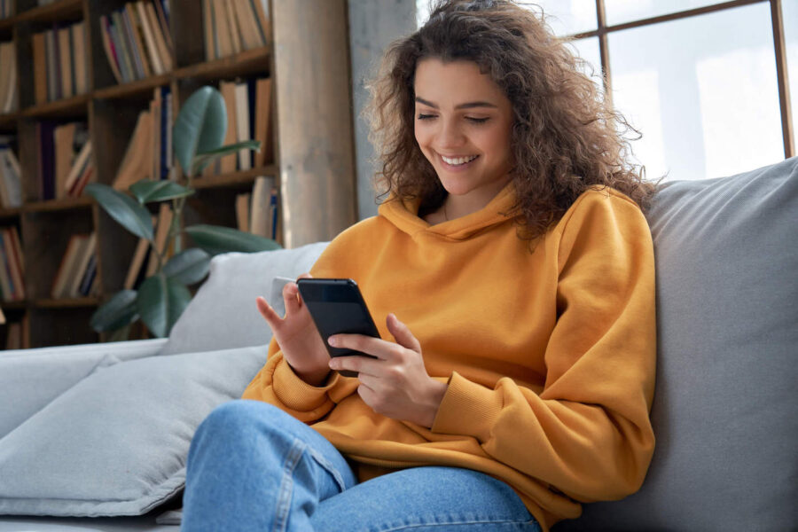 A young woman wearing an orange sweater is sitting on the couch while using her phone.