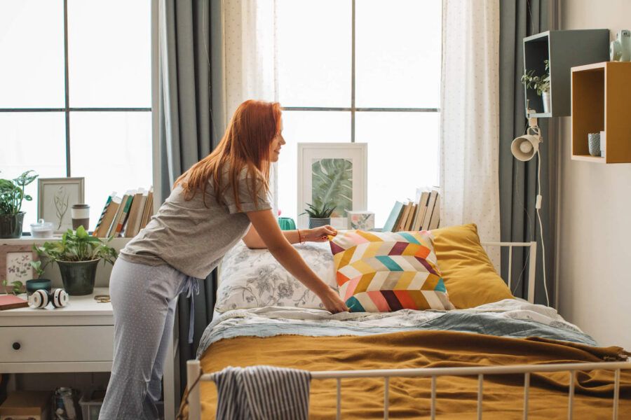 A young woman with red hair adjusts a pillow on her bed.
