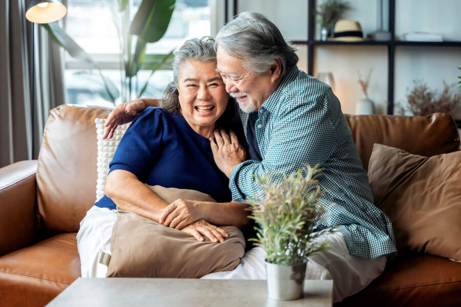 An elderly couple sitting on a brown couch are hugging and laughing with each other.