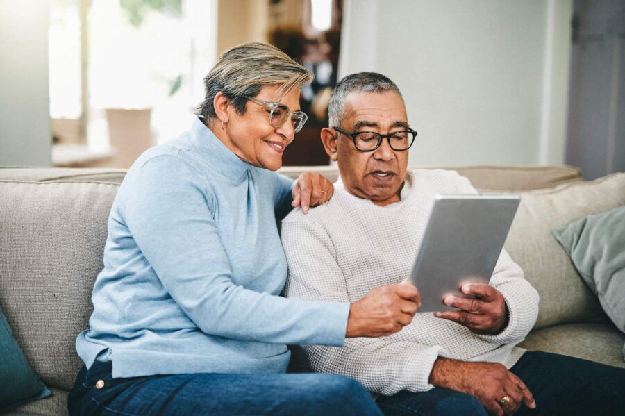 An elderly couple sits on the couch together while looking a tablet screen.