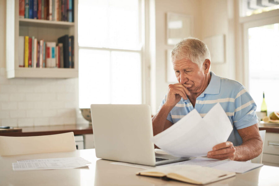 An elderly man looks at his laptop as he frowns while holding documents in his hands.