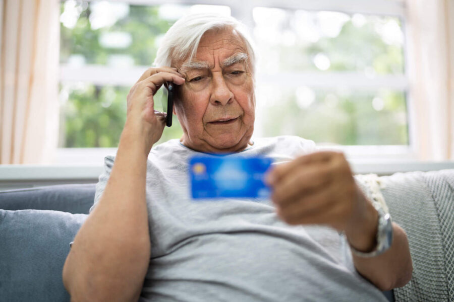 An elderly man sits on the couch while talking on the phone and holding his credit card.
