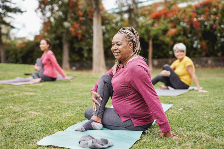 An elderly woman smiles while doing yoga at the park with two woman behind her.
