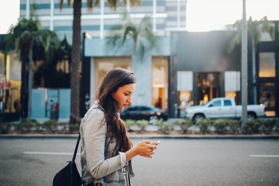 A woman looks at her phone while walking down the sidewalk outside.