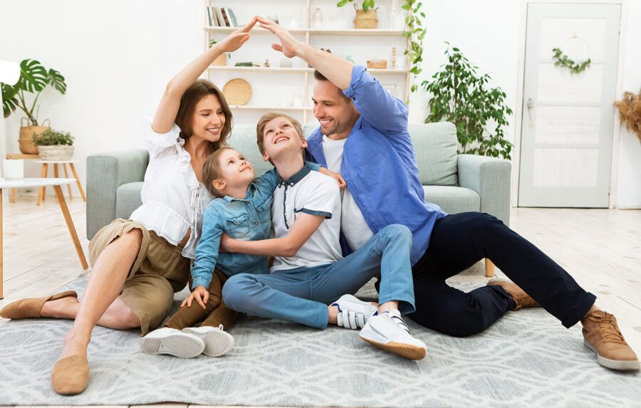 A family of four sit on the ground of their living room as the mom and dad connect their hands over their children.