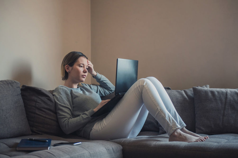 Woman lying down on sofa and desperately looking for a job on her laptop