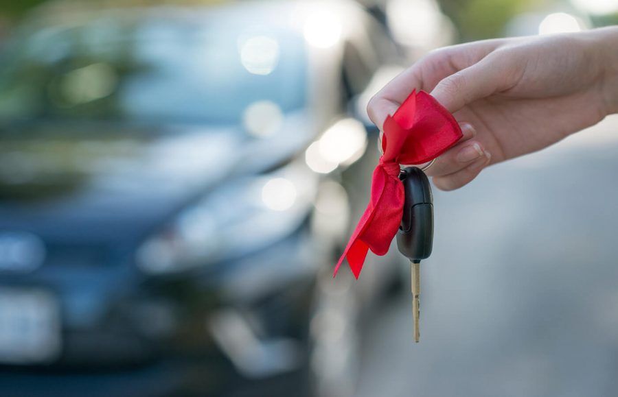 A hand holding a car key with a red bow tied to the key ring, held in front of an out-of-focus sedan car in the background.