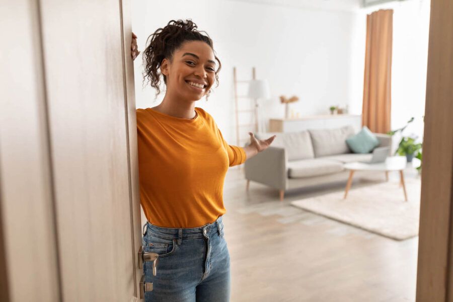 Cheerful African American woman opening door and gesturing inside house.