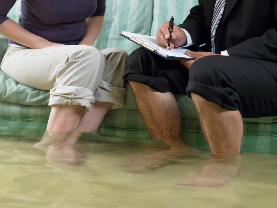 A closeup photo of a homeowner's and agent's feet and legs underwater as they sit on a couch in a flooded home while the agent writes on a document with their pants rolled up.