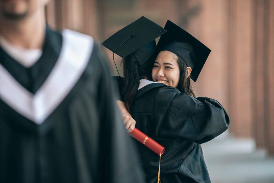 Two college students are hugging each other while wearing their graduation gowns.