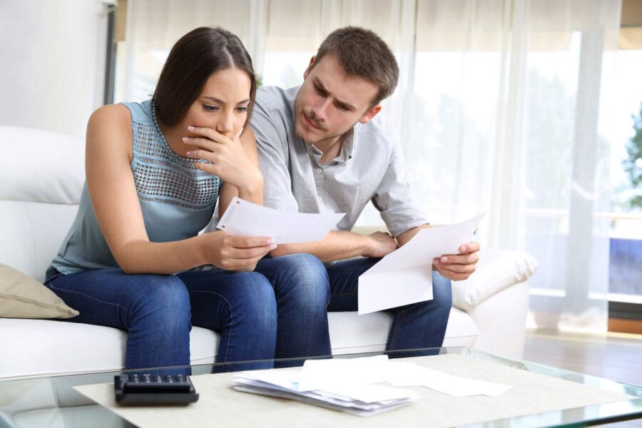 A couple sitting on the couch frown together while holding documents.