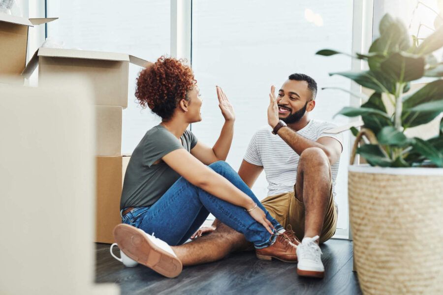 A couple sits on the floor and high-five each other while being surrounded with boxes in their new home.