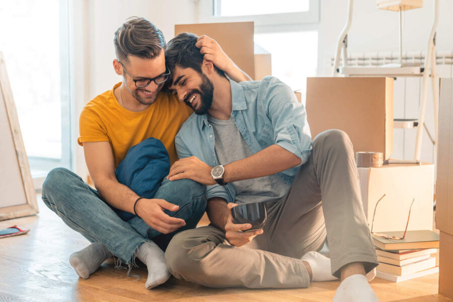A couple laugh and embrace each other on the floor of their new home with boxes in the background.