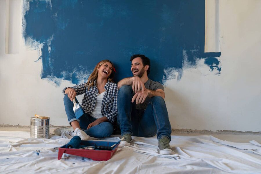 A couple sitting down on the floor laugh together while they are painting the wall blue.