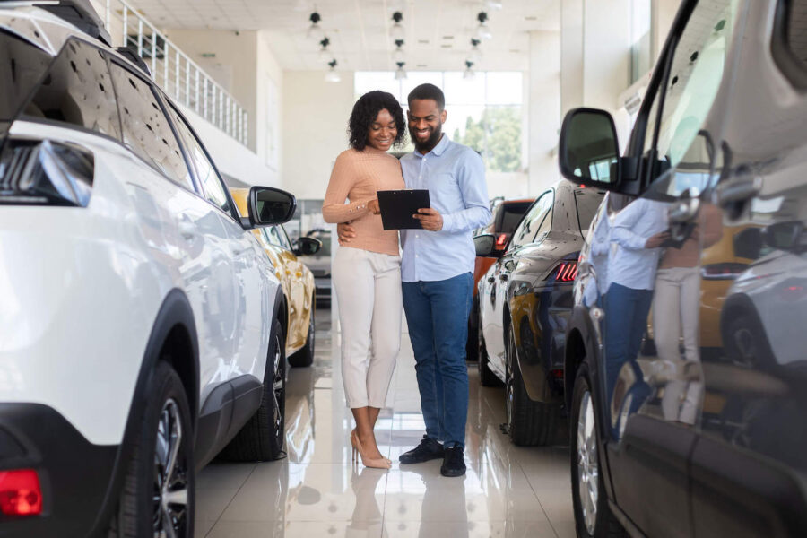 A couple smiles while looking at a document at the car dealership while surrounded by new cars.
