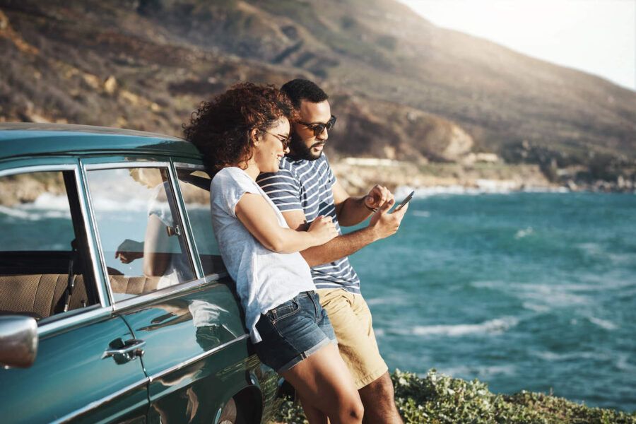 A couple look at their phone together while leaning on their car with the ocean in the background.