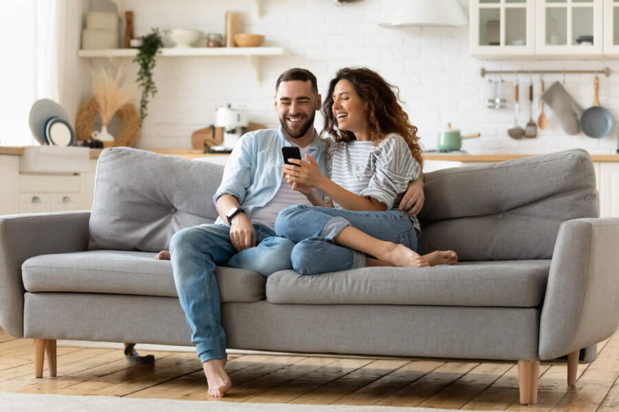 A couple sit on the couch together and smile while they look at a phone.