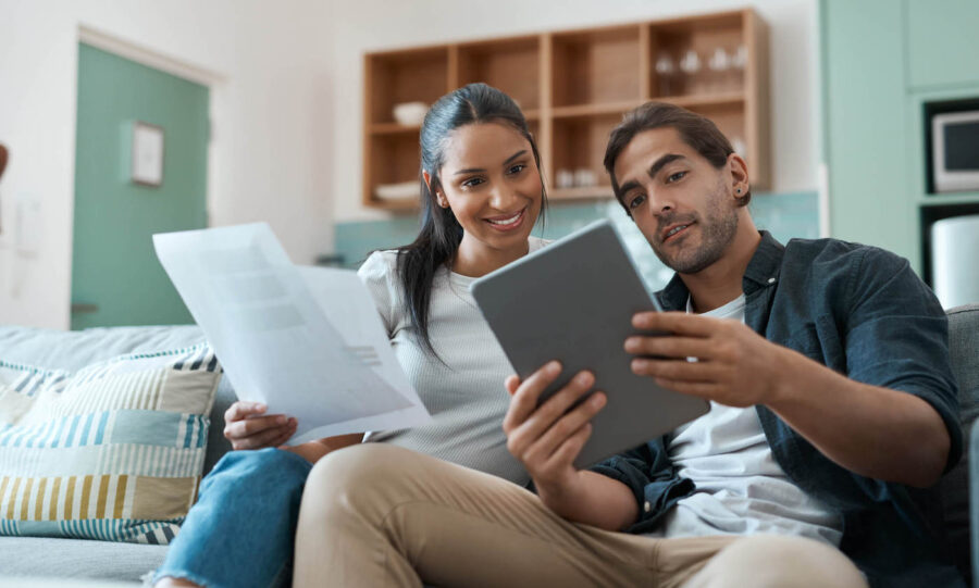 A couple sit together on the couch while looking at a tablet and holding documents.