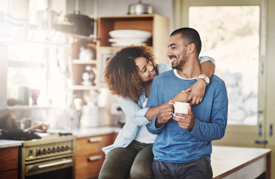 A couple smile and embrace each other in the kitchen while the man holds a coffee cup.