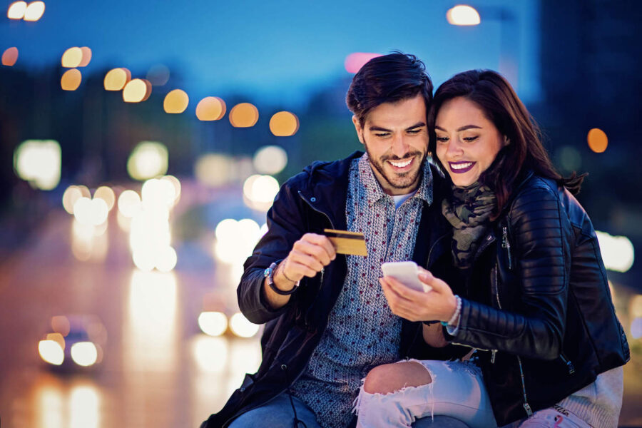 A couple outside smile at a phone together while the man holds a credit card.