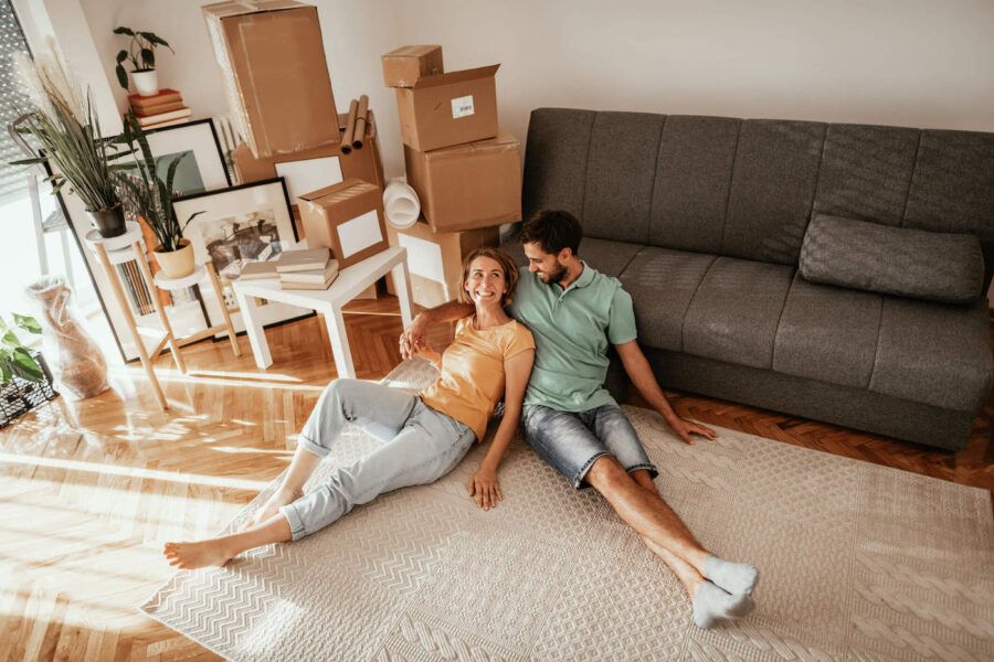 A couple on the floor smile together while inside of their new home with boxes in the living room.