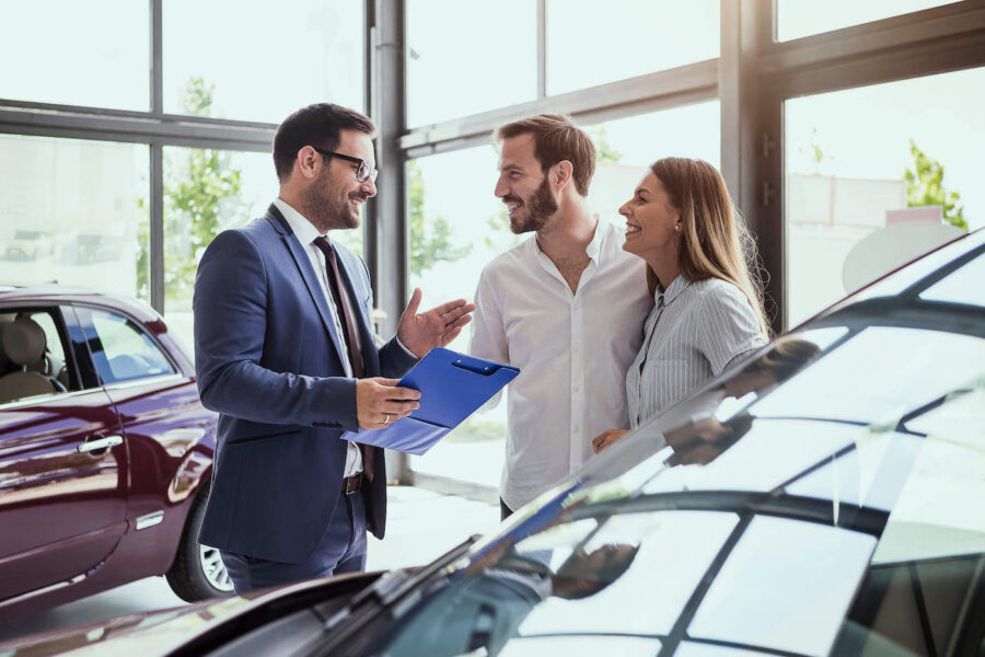 A couple smiles at a car salesman wearing a blue suit at the car dealership.