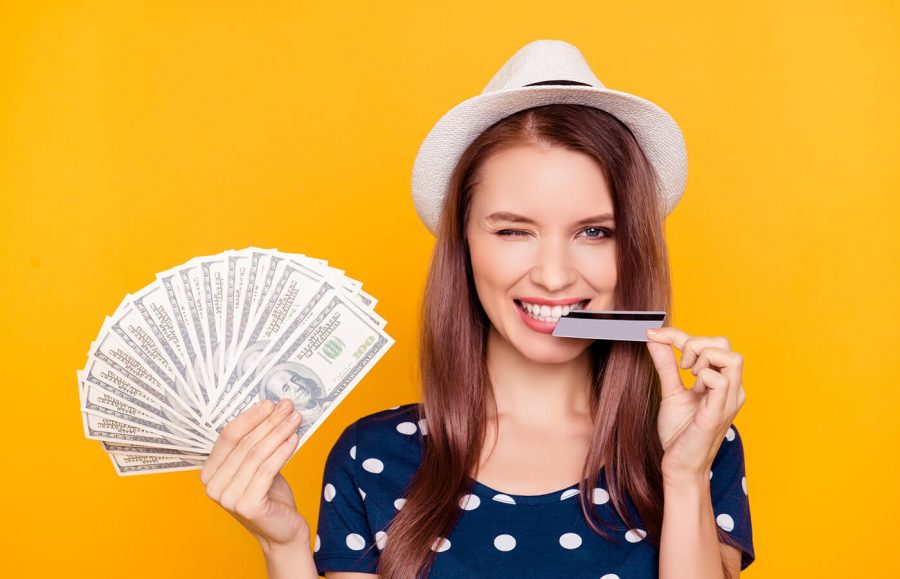A woman biting her credit card while holding cash in her other hand with a yellow wall in the background.