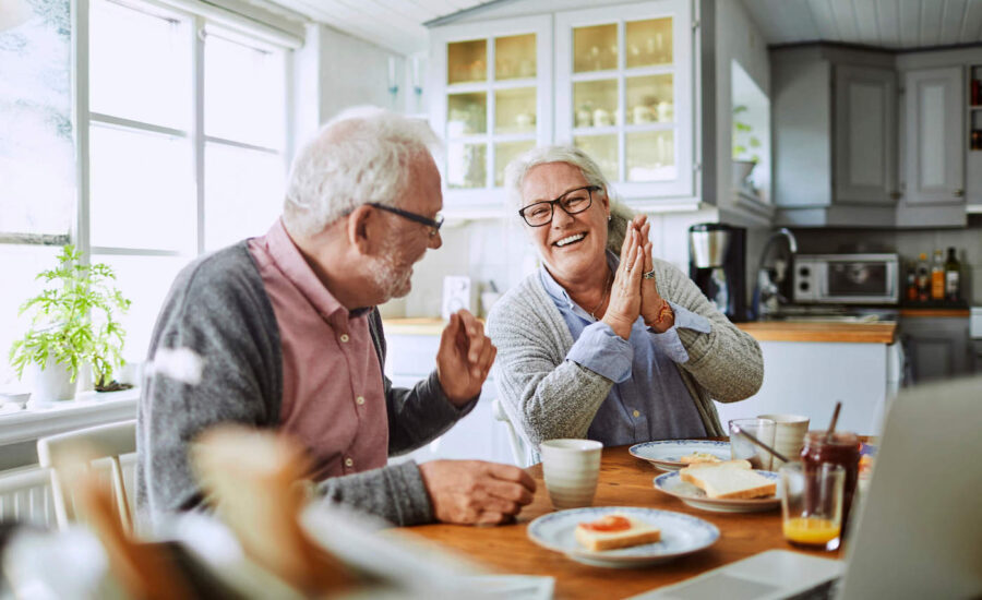 An elderly couple laugh together while eating breakfast at the kitchen table.
