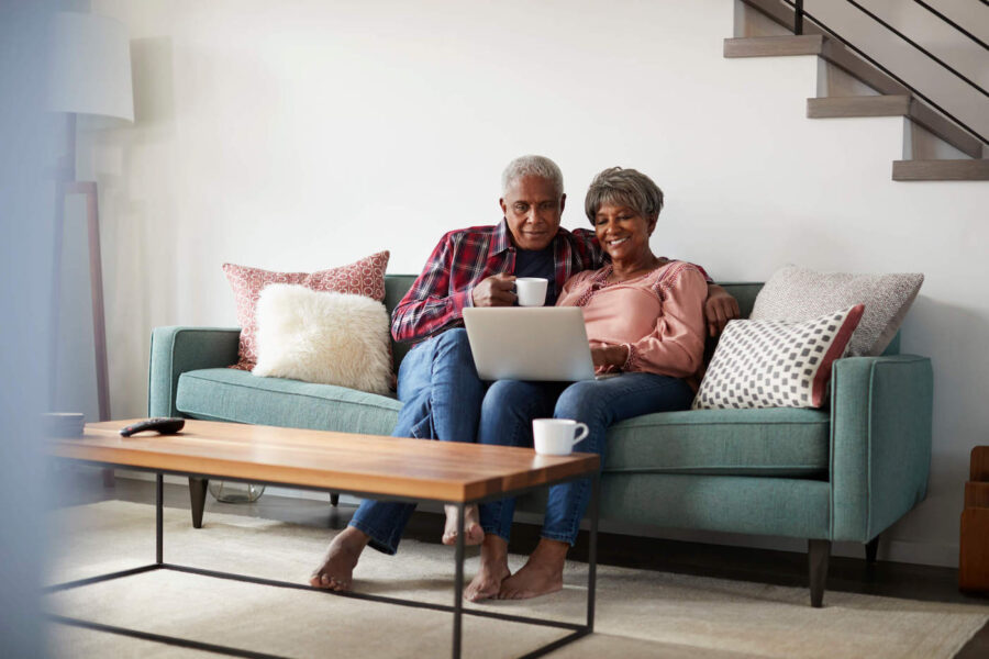An elderly couple look at a laptop computer together while sitting on the couch at home.