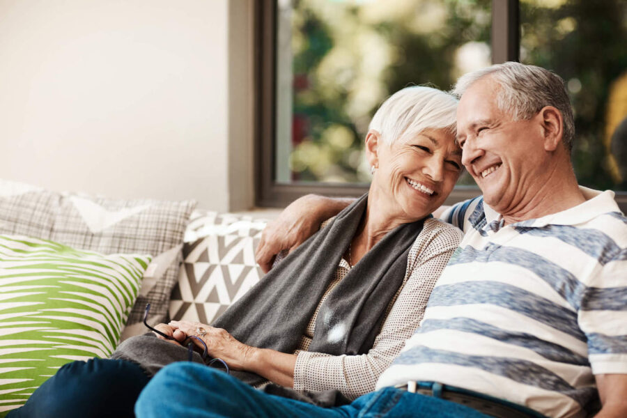 An elderly couple smile and embrace each other while sitting on the couch.