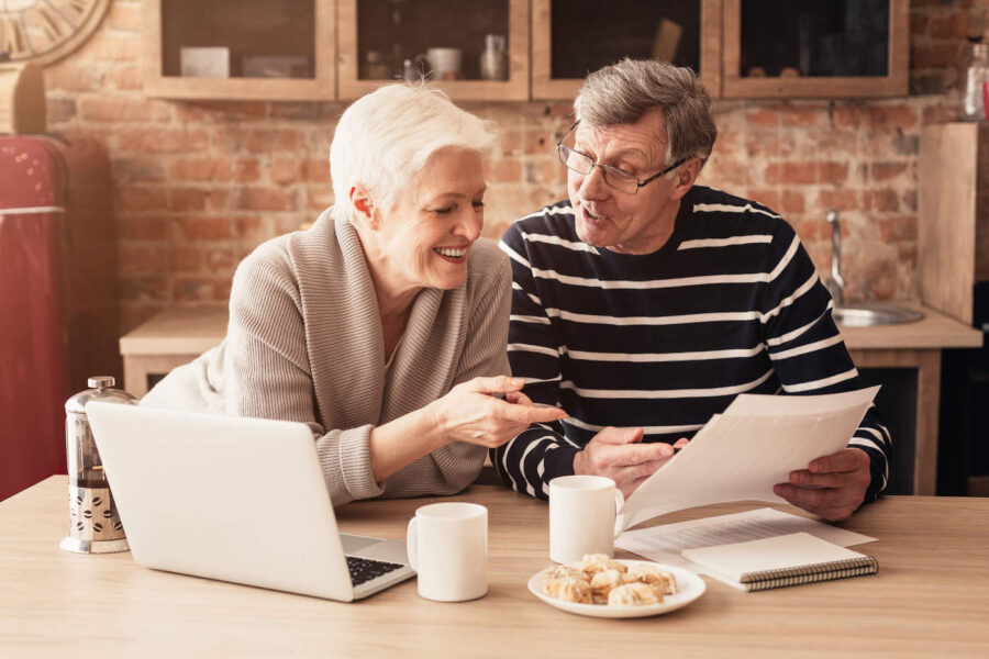 An elderly couple smiles as they look at a document while their laptop, coffee and breakfast sits on the kitchen table.