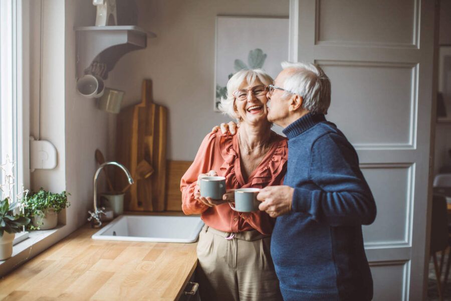 An elderly man kisses his elderly wife in the cheek while they are holding their coffee cups in the kitchen.