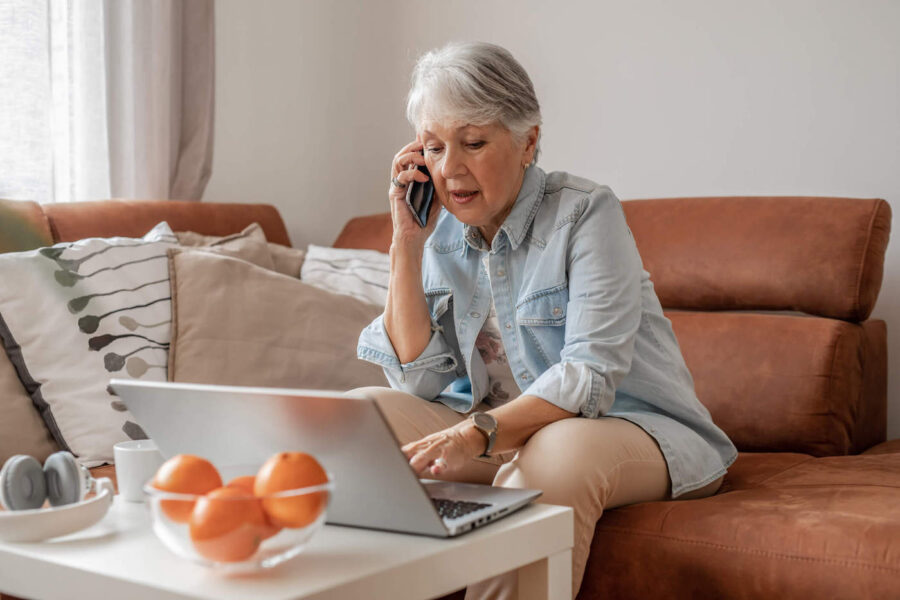 An elderly woman sitting down on her couch uses her laptop while talking on the phone.