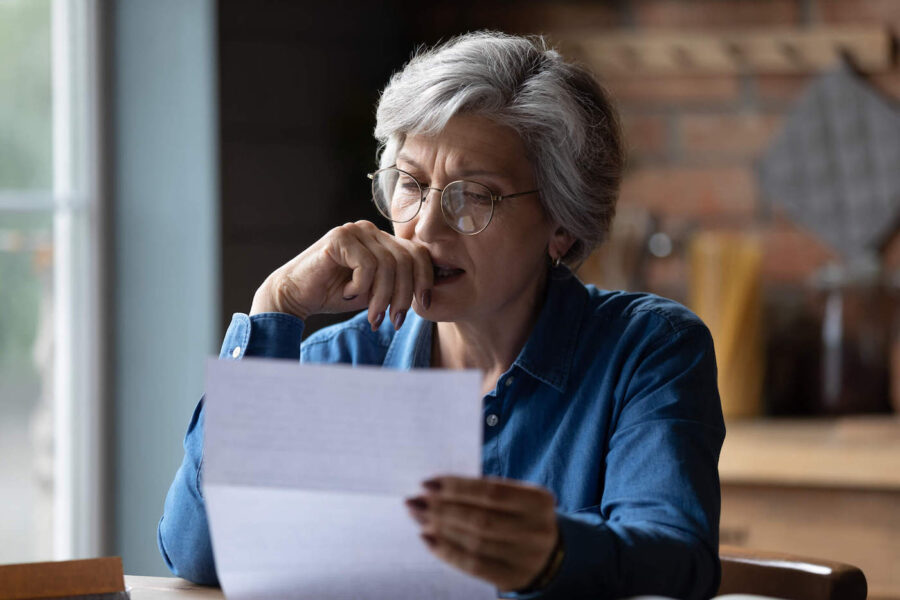 An elderly woman wearing glasses has her hand to her mouth as she looks at a document.