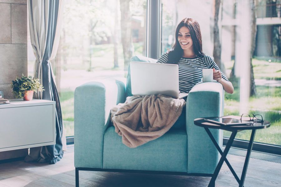 Beautiful young smiling woman working on laptop and drinking coffee while sitting in a big comfortable chair at home
