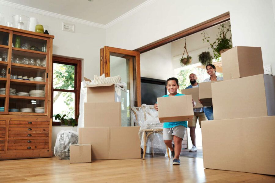 A family of three are smiling while carrying brown moving boxes as they move into their new living room that has wood flooring, a wooden cabinet filled with kitchen items, and furniture wrapped in plastic.
