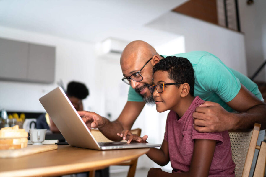 A father points at a laptop screen while his son uses the laptop on the kitchen table.