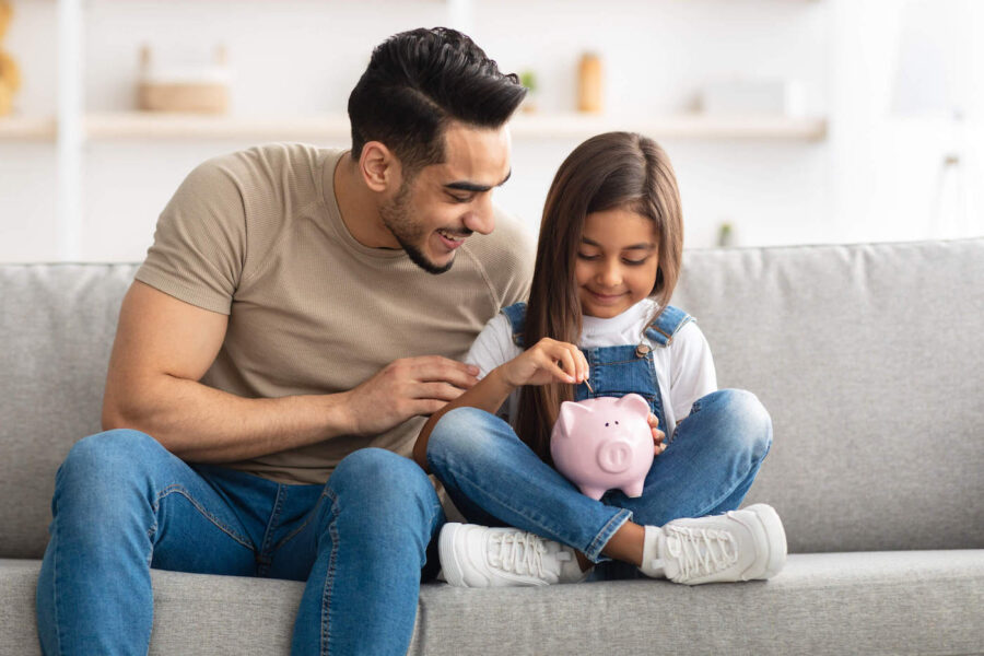 Father and daughter are smiling on the couch while they are putting a coin in a pink piggybank.