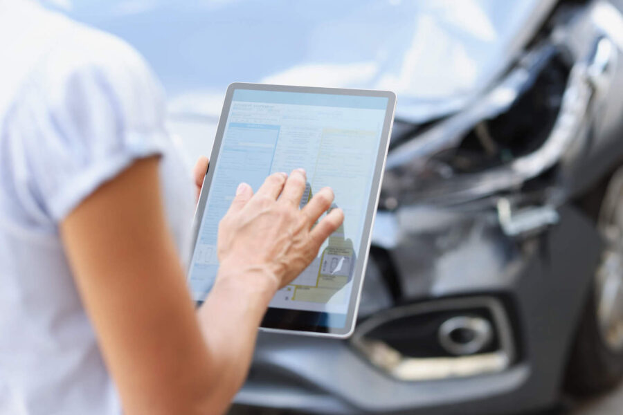 Female agent filling out insurance claim on digital tablet against background of broken car closeup.