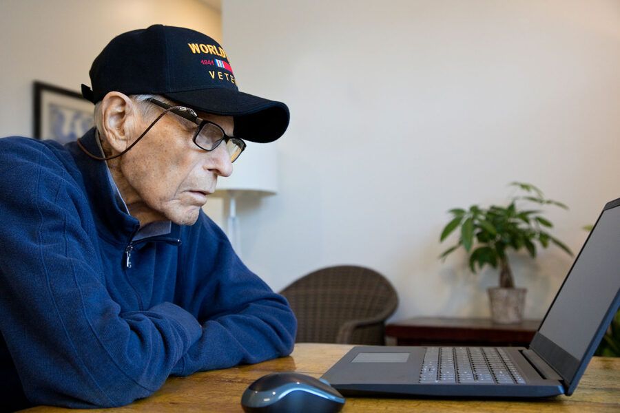 An elderly man wearing a veteran hat looks at his laptop screen while his arms are crossed.