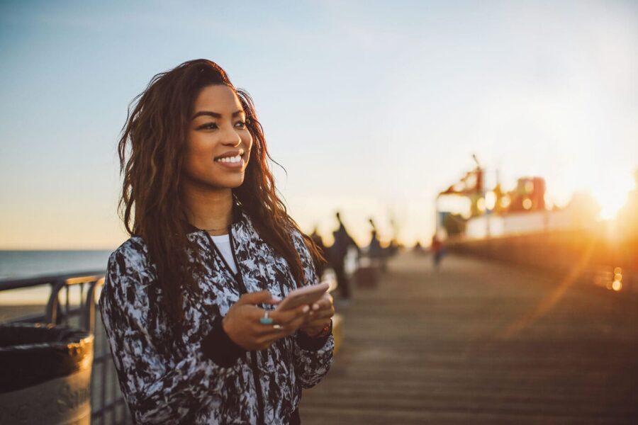 A smiling woman holding her phone and standing on a pier.