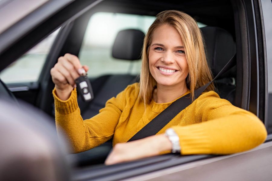 A smiling woman sitting in the driver's seat of her new car, triumphantly holding up the keys and resting her elbow on the car door.