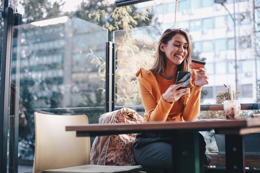 A young woman at a restaurant smiles at her credit card while holding her phone in the patio area.