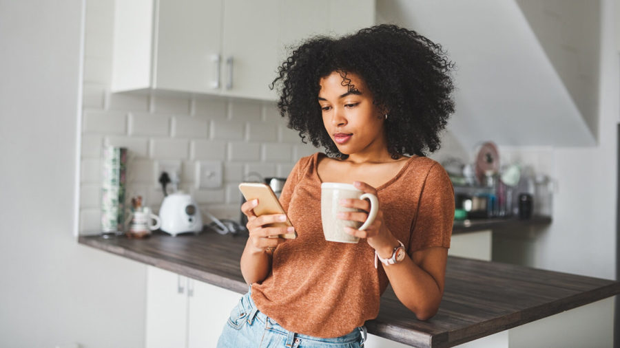 A young woman looks at her phone while holding a coffee cup in her other hand.