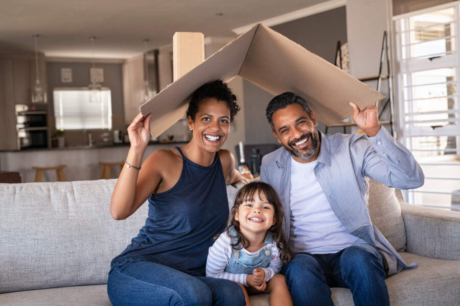family of three sitting on a couch holding a cardboard roof above their heads