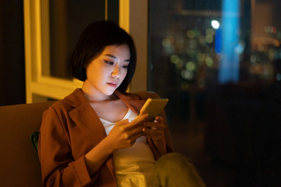 A woman sitting on the couch looks at her phone in a dark room.