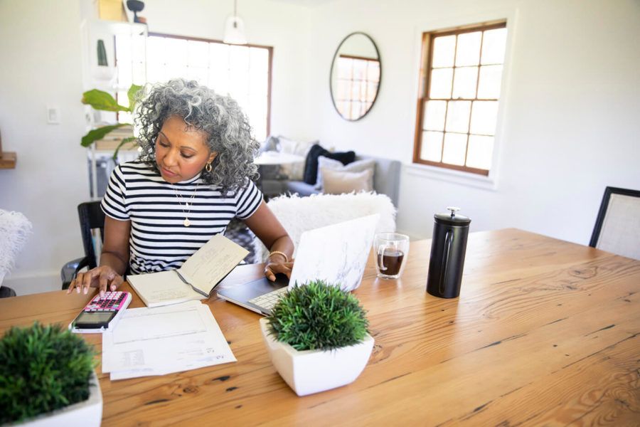 A beautiful black woman with white curly hair works at home on her laptop with her coffee and a calculator